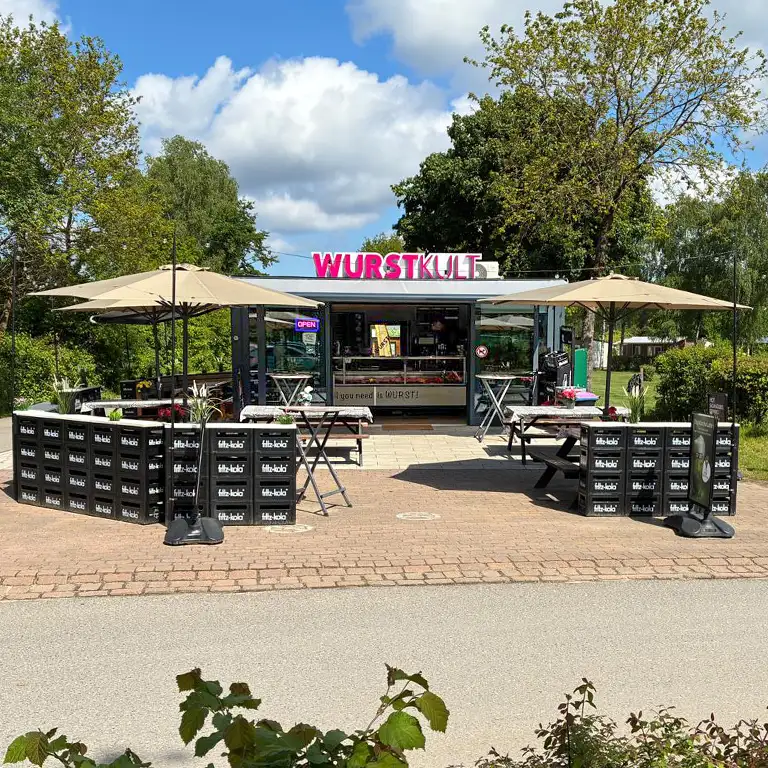 Imbissstand im Südsee-Camp mit der Aufschrift ‚Der Küstenhimmel wird jetzt zu WURSTKULT‘ vor einer Wursttheke mit Fleischwaren und Slogan ‚All you need is WURST‘.